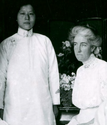 Black and white photo. Young woman Tien Fuh Wu stands in a high-collared white blouse behind and to the left of a mature woman (Donaldina Cameron) seated in a parlor at the Presbyterian Mission House, circa 1910s.