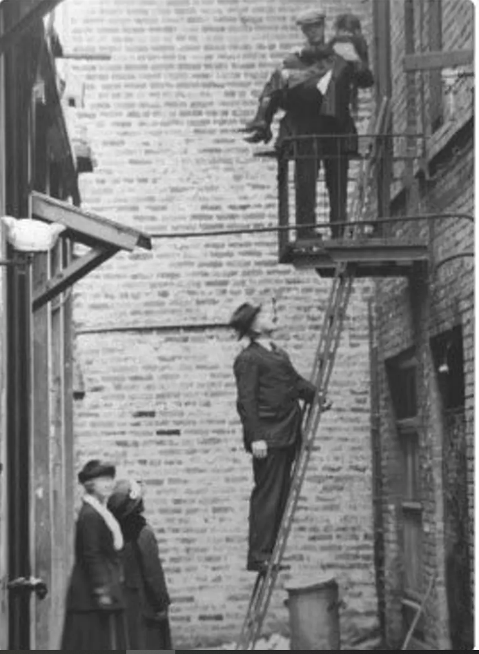 Undated black-and-white photo of a rescue in a narrow Chinatown alley. A police officer on a fire escape cradles a young girl; below, a man steadies a ladder. At ground level, Donaldina Cameron stands at bottom left beside an unidentified woman, both looking up.